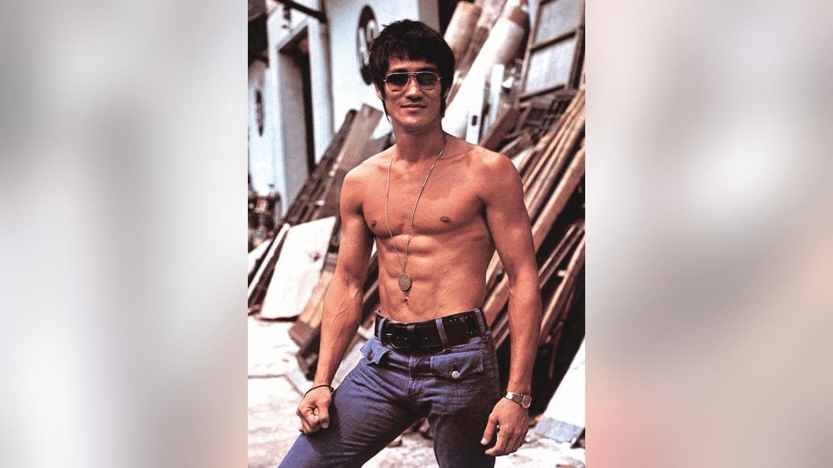 Bruce sem camisa, usando colar medalhão, óculos escuros, sorrindo, em pé diante da construção do cenário.