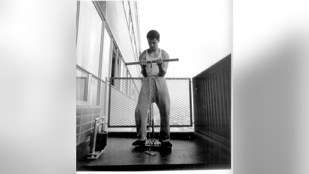 Bruce wearing tank top, sweats, demo of isometric reverse curl, with weight, on patio of apartment high rise.