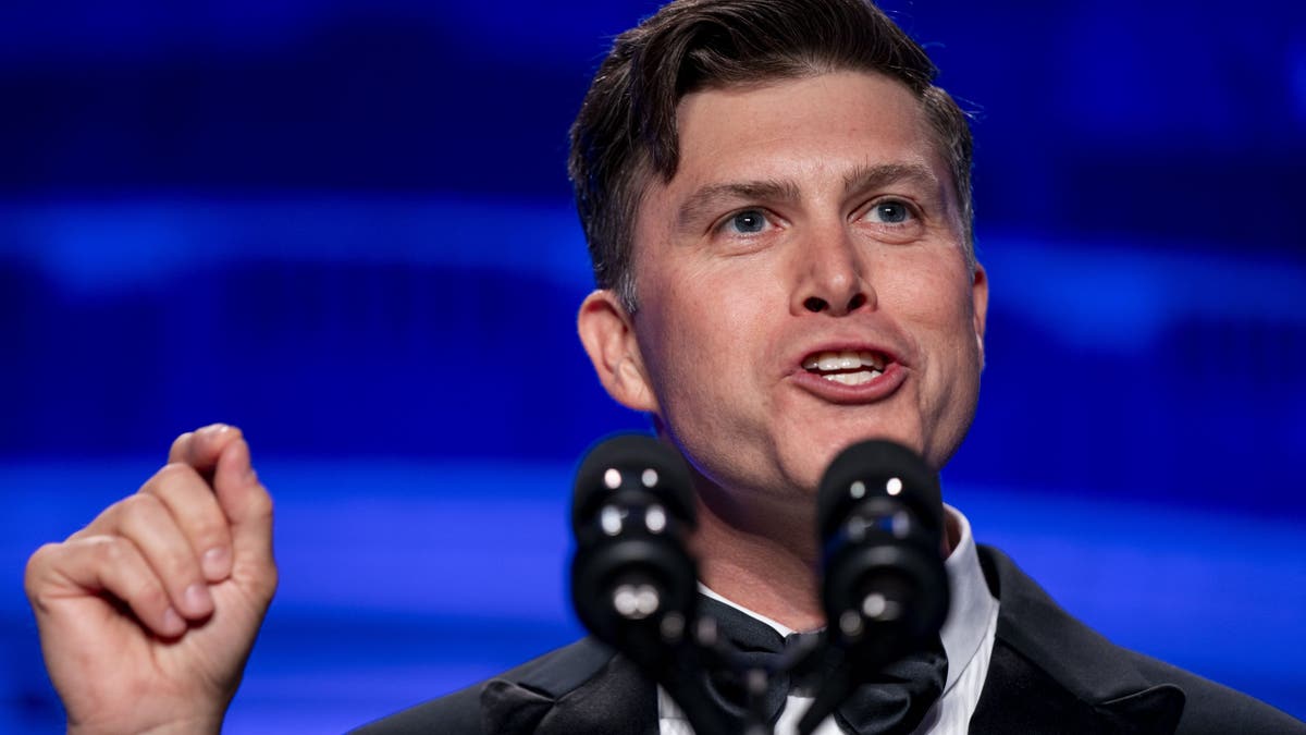 Comedian speaking at podium on stage with microphone, blue background.