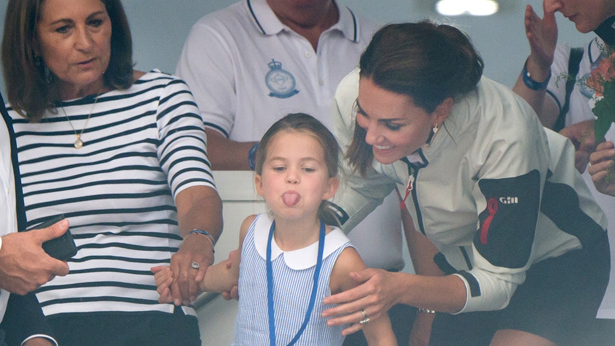 Carole Middleton segurando a mão da princesa Charlotte enquanto mostra a língua para uma multidão
