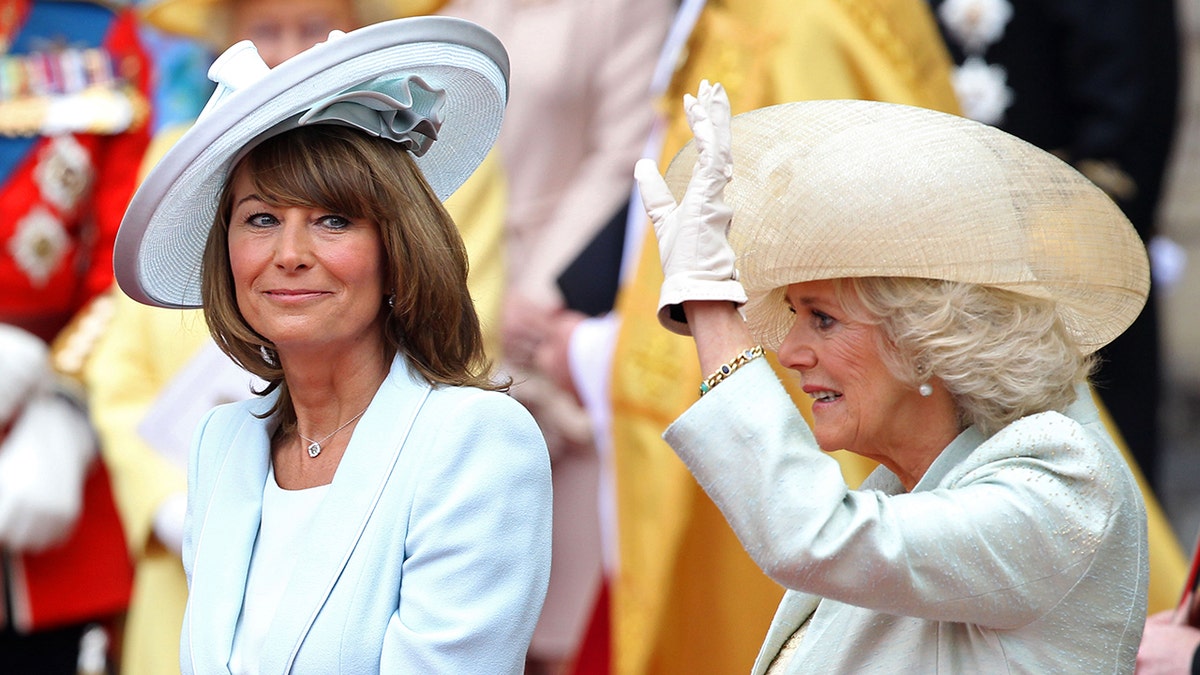 Carole Middleton sentada ao lado da Rainha Camilla, que está acenando para uma multidão