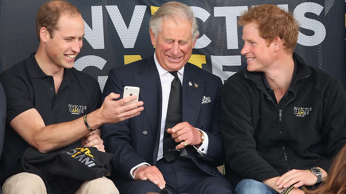 Prince William shows his cell phone to a laughing King Charles as Prince Harry smiles