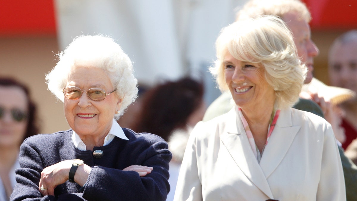 Queen Elizabeth II smiling with her arms cross next to Camilla who is also smiling