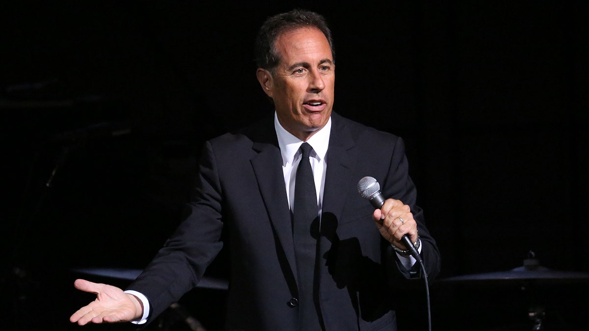Jerry Seinfeld in a black suit and tie holds a microphone and performs on stage