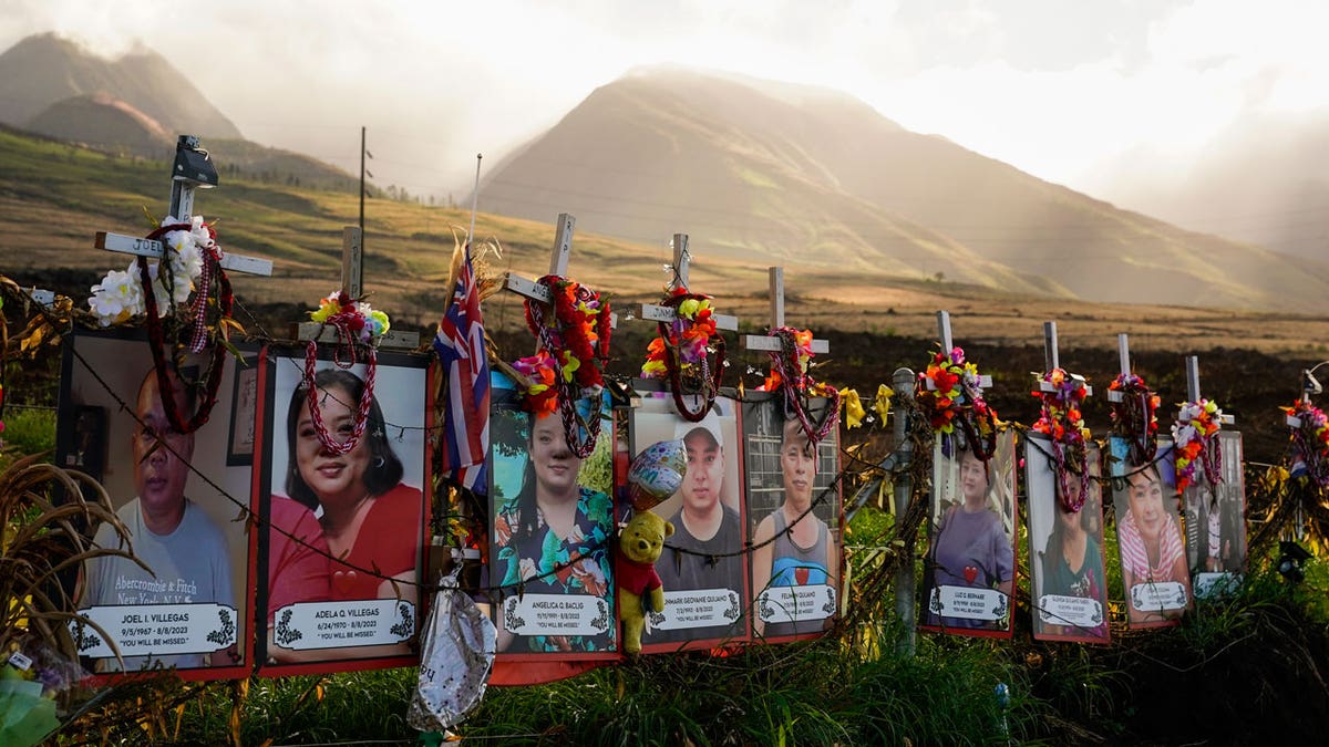 Photos of victims are displayed under white crosses at a memorial for victims of the August 2023 wildfire in front of a scenic mountain view