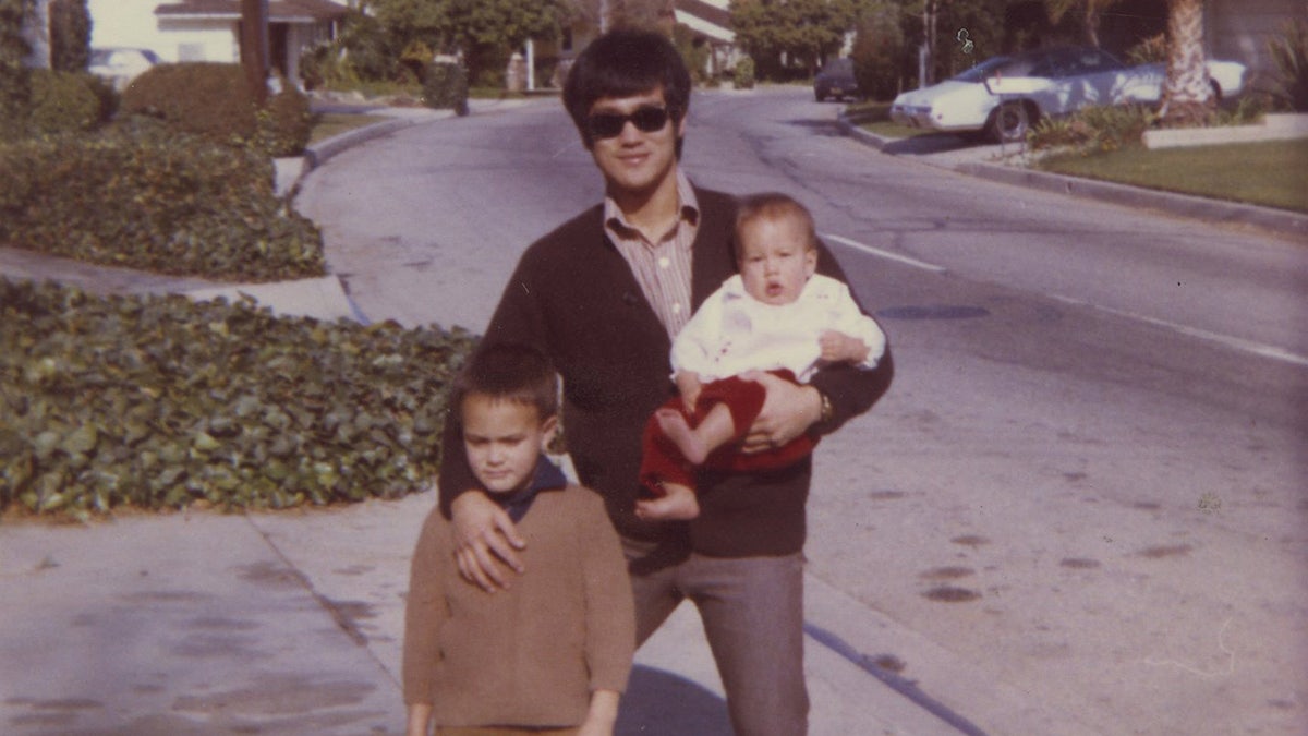 Bruce in dark sweater, brown shirt, sunglasses, smiling in driveway of home holding Shannon with hand on Brandons chest. Family, portrait, houses, street, and palms in background.