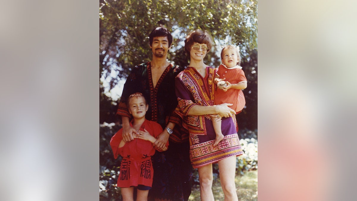 Family portrait, Bruce, Brandon, Linda, Shannon. Bruce is wearing a dark kaftan, beard, smiling, hand on Brandon, while Linda holds Shannon. Tree and grass in background.