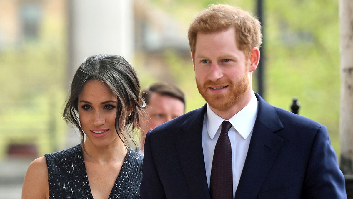 Meghan Markle wearing a black sparkling v-neck dress next to Prince Harry in a suit and dark tie