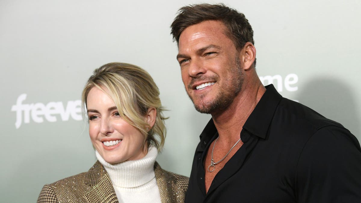 Alan Ritchson and wife Catherine smile on carpet.