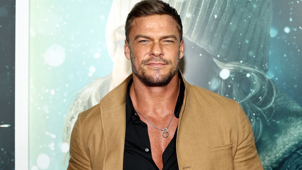 Alan Ritchson in a black button up and tan jacket smiles on the carpet.