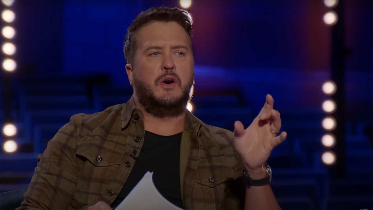 Luke Bryan in a brown plaid shirt over a black shirt puts his hand up and holds papers on "American Idol"