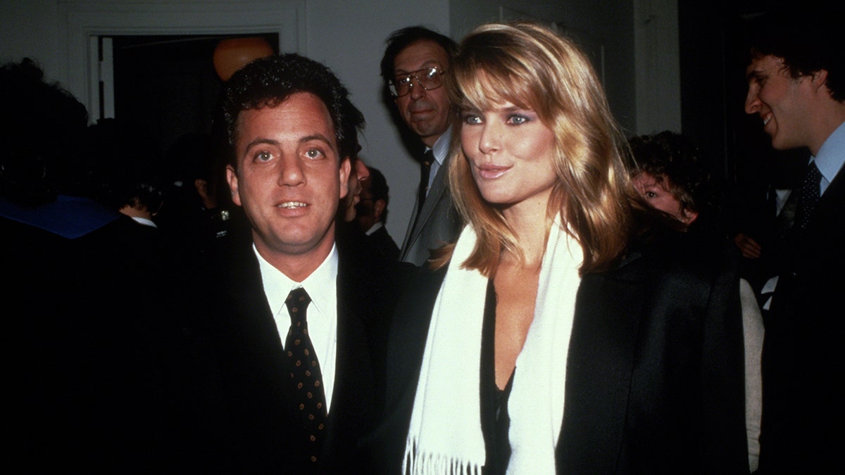 Billy Joel in a suit stands next to Christie Brinkley in a dark coat and white scarf