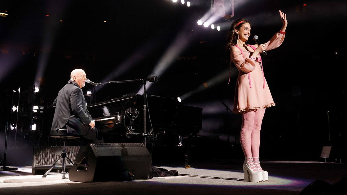 Alexa Ray Joel at the front of the stage in a pink dress and tights with her father Billy Joel behind her at the piano