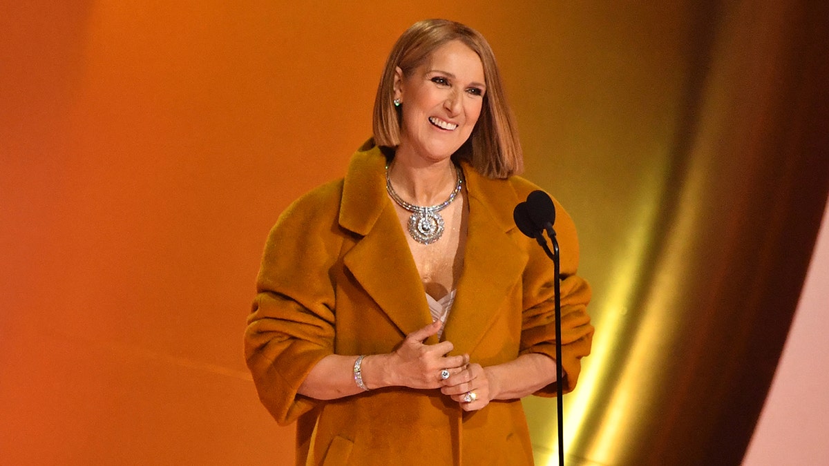 Céline Dion in a large orange coat smiles on stage at the Grammys