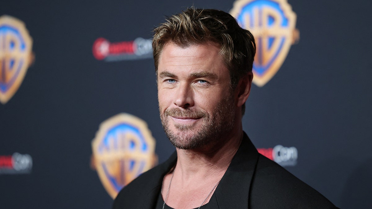 Chris Hemsworth sorri suavemente no tapete em uma jaqueta preta