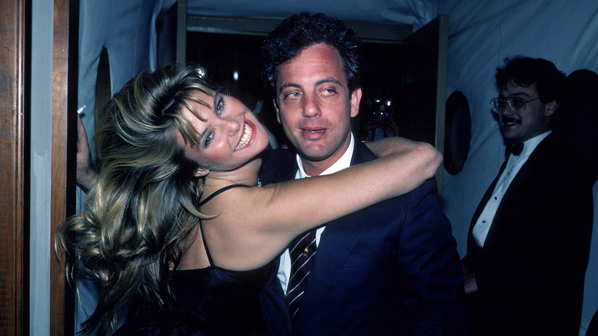 Christie Brinkley in a black dress wraps her arm around Billy Joel looking away from the camera