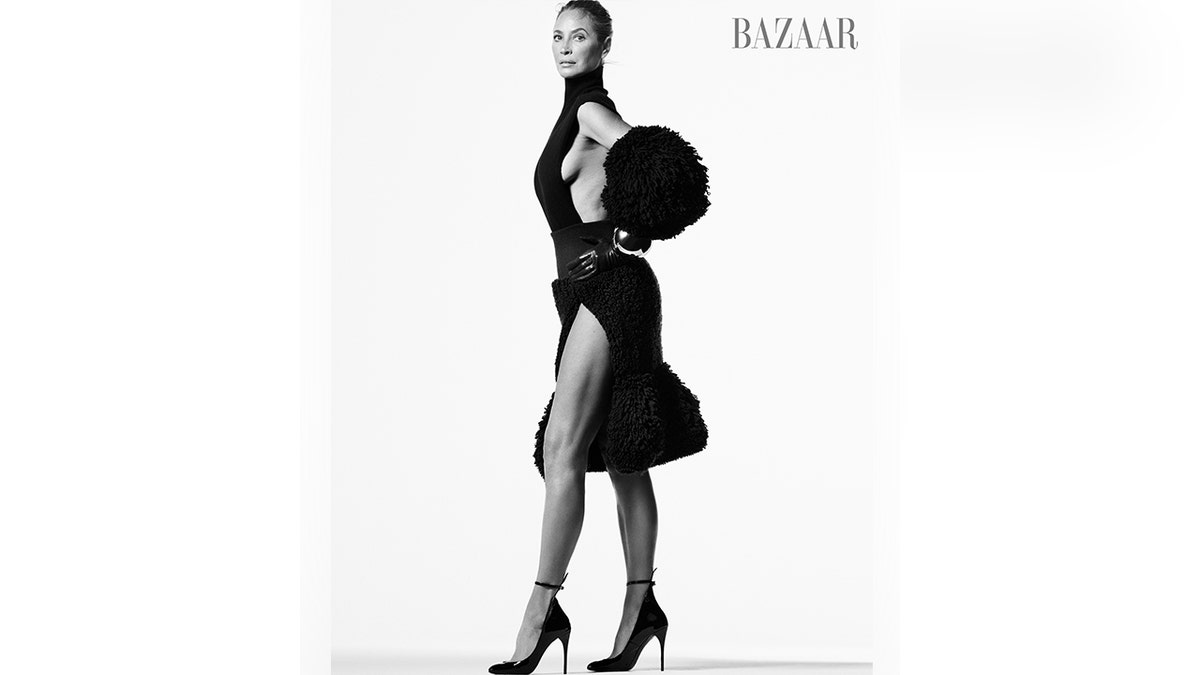 Christy Turlington in a black and white photo for Harper's Bazaar stands in a black dress with a slit with a poofy bottom and elbow pads/poofs