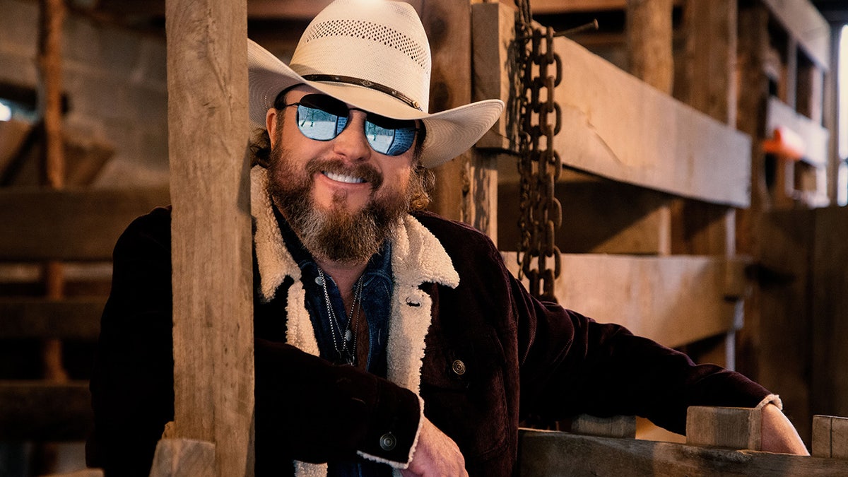 Musician Colt Ford wears a cowboy chapeau and sunglasses successful a barn.