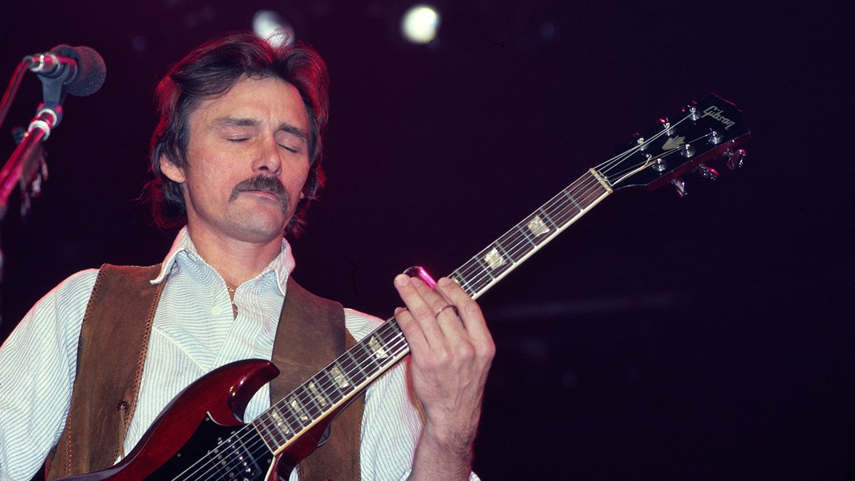 Dickey Betts in a brown vest strums the guitar