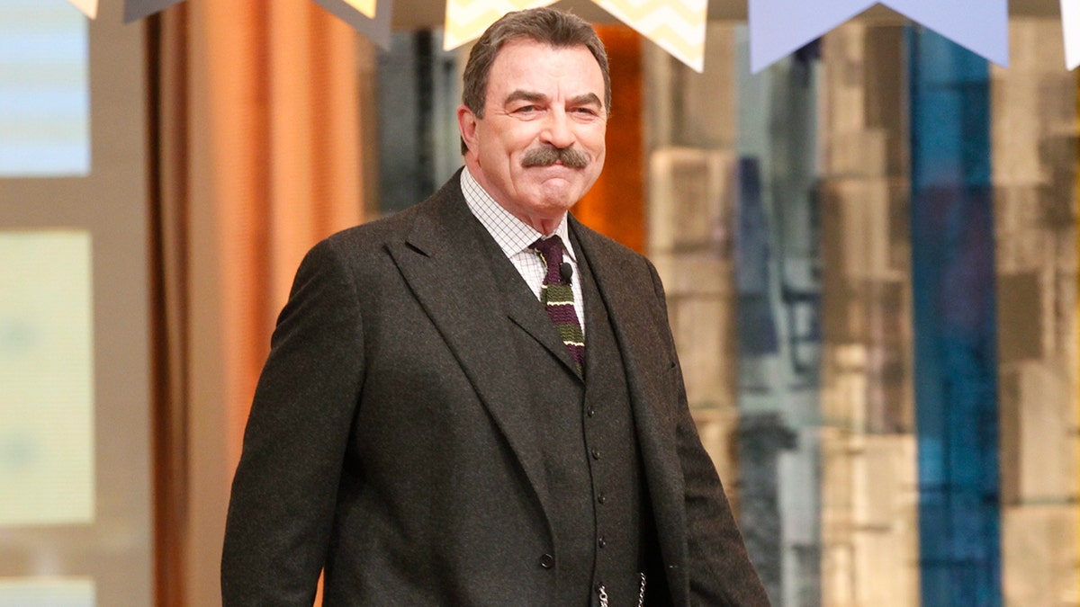 Tom Selleck in a black suit with a slight smile looks at camera
