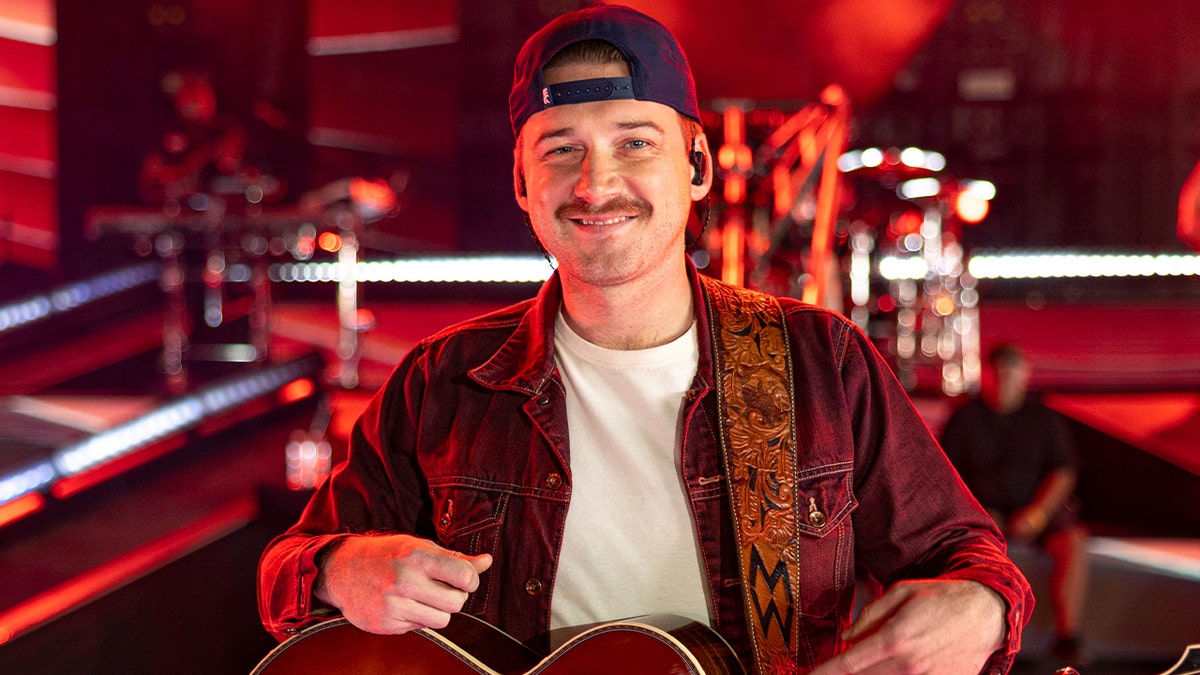Morgan Wallen smiling in a denim jacket and backwards hat, holding his guitar