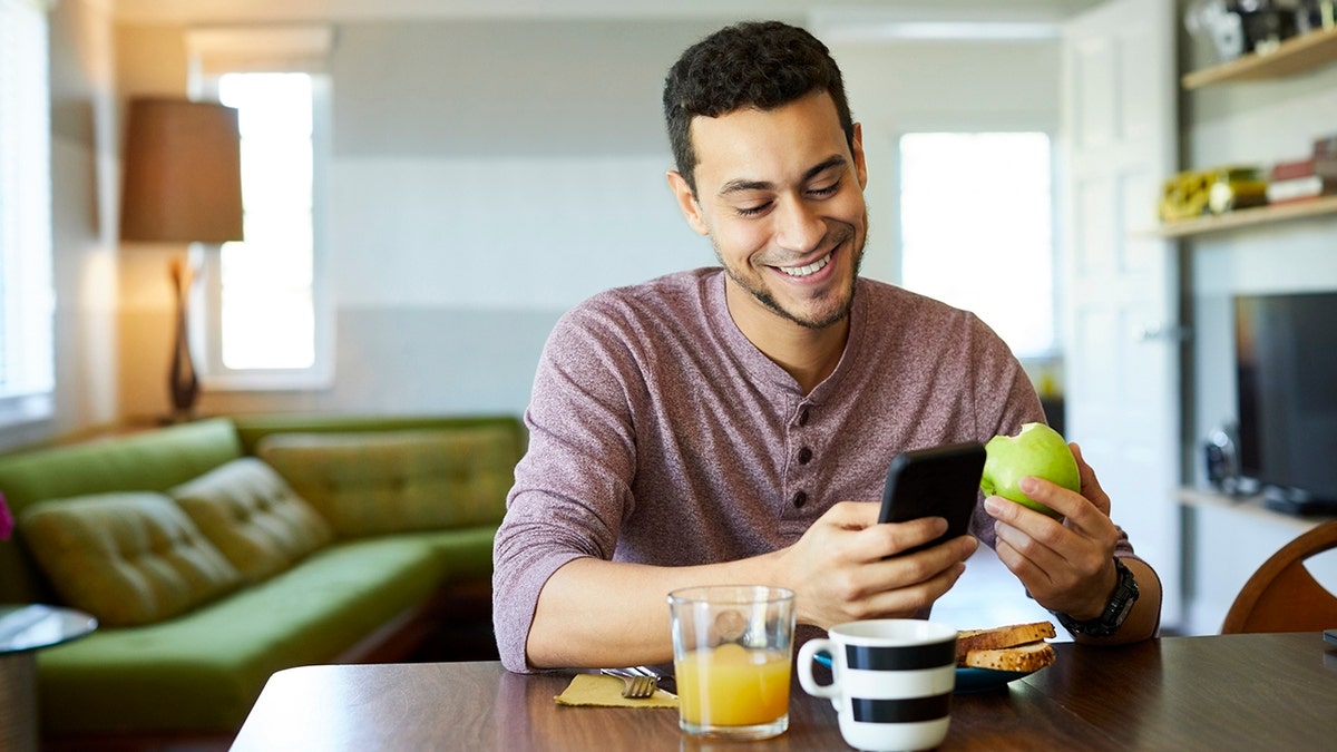 man eats an apple while scrolling on his phone in front of breakfast