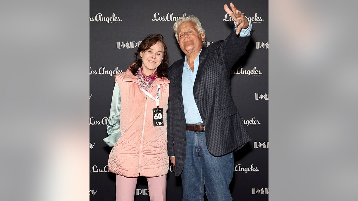 Mavis Leno in a peach colored vest and light turquoise blouse and pink pants smiles with Jay Leno in a black suit, jeans and blue shirt on the carpet