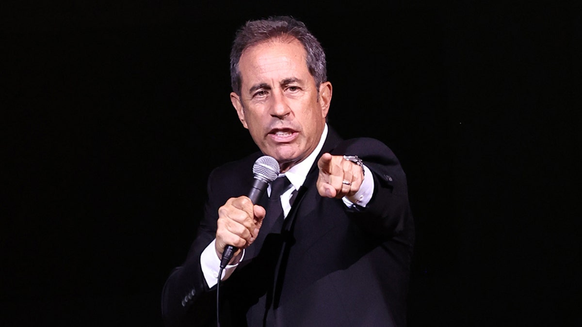 Jerry Seinfeld in a black suit on stage points directly out towards the crowd