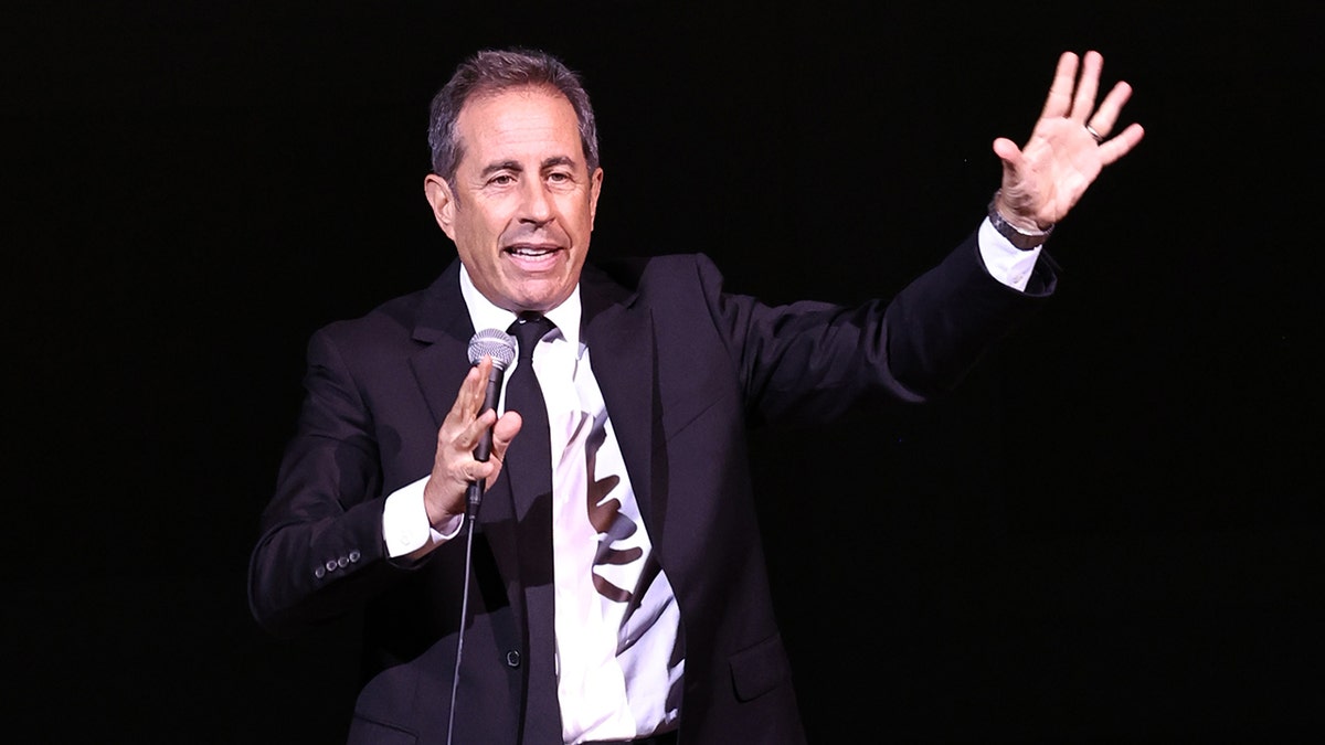Jerry Seinfeld with his hand up in the air speaking into a microphone on stage