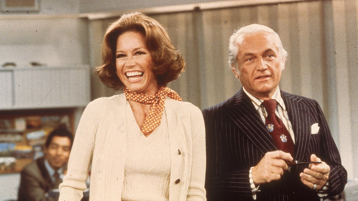 Mary Tyler Moore in a white sweater laughs on set with Ted Knight in an episode of "The Mary Tyler Moore Show"