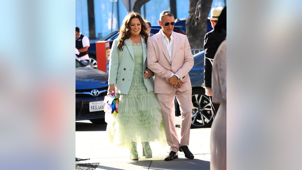Melissa McCarthy in a mint green tulle dress with a matching blazer walks with director Adam Shankman in a light pink suit