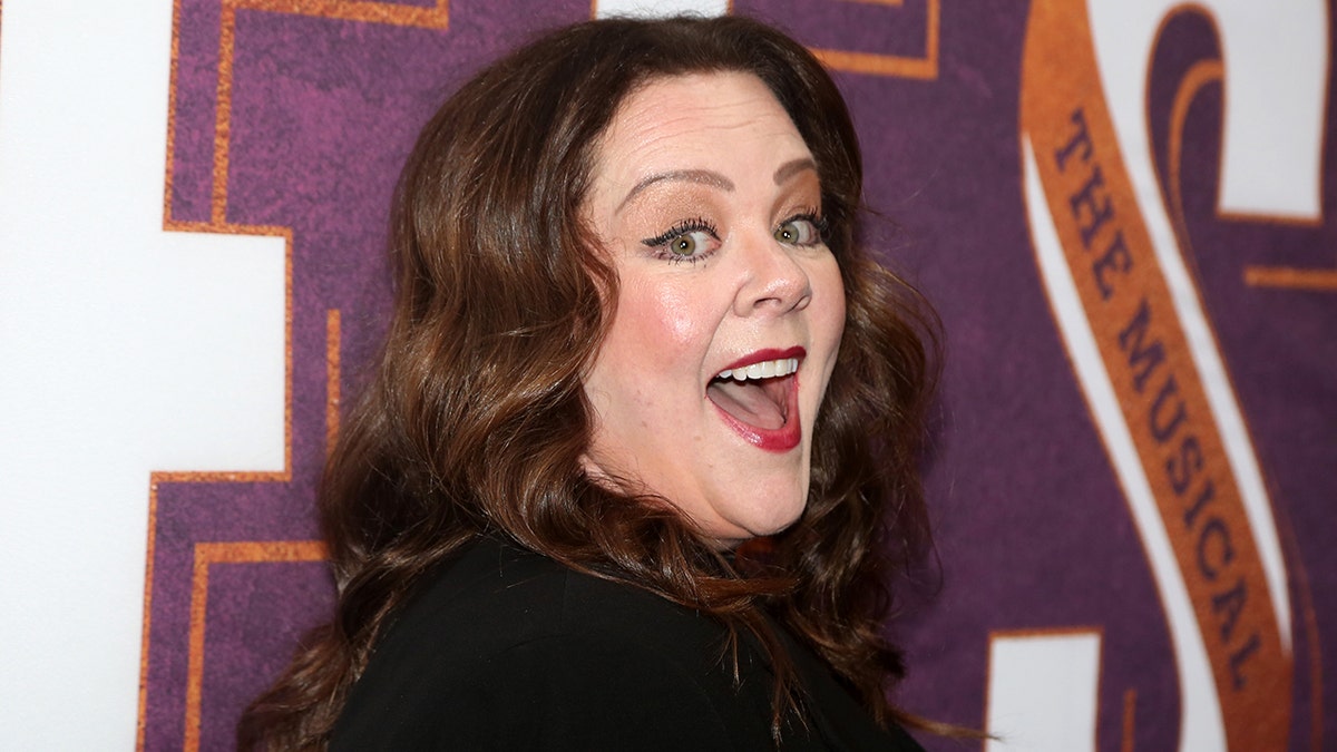 Melissa McCarthy looks partially over her shoulder in a black outfit on the carpet with a wide open smile