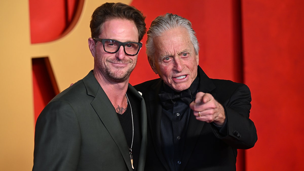 Cameron Douglas in a green jacket and black frame glasses soft smiles on the carpet with father Michael Douglas in a black suit who points at the cameras