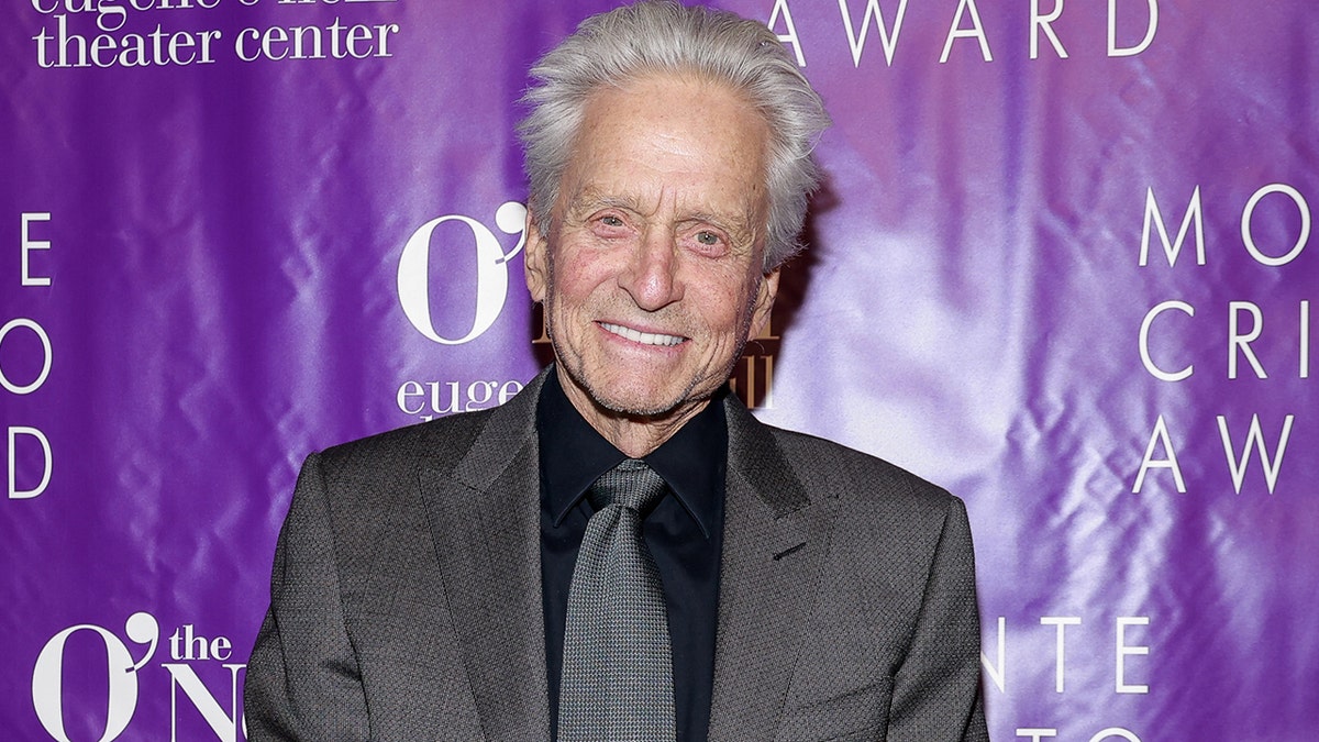 Michael Douglas smiling behind a purple backdrop on a red carpet