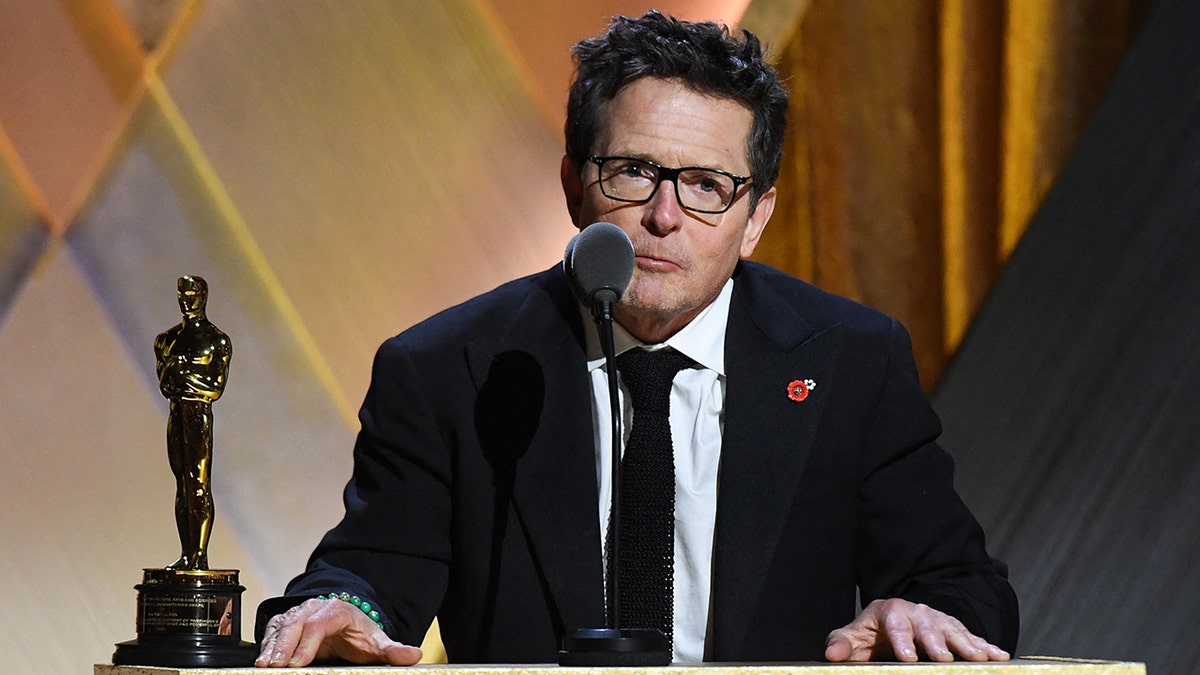 Michael J. Fox in a black suit stands behind the podium to accept his honorary Academy Award