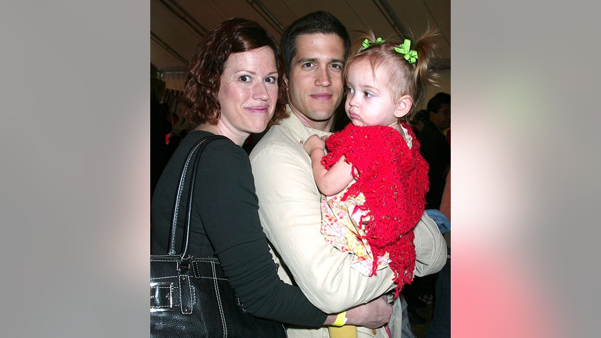 Molly RIngwald in black hugs her boyfriend Panio Gianopoulos in a cream sweater who holds their daughter Mathilda in a red dress