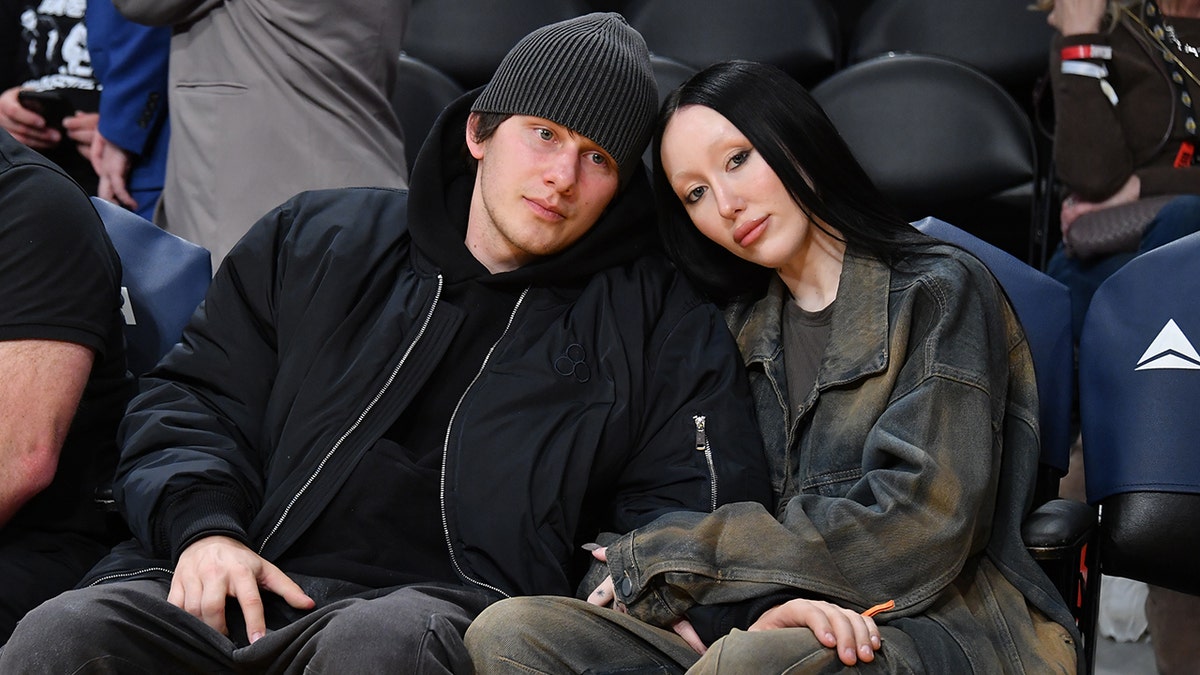 Noah Cyrus, con una chaqueta de cuero oscura, apoya la cabeza en su prometido Pinkus, sentado junto a la cancha en el partido de los Lakers