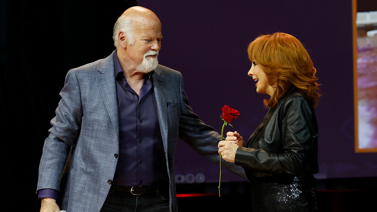 Rex Linn in a blue jacket gives Reba McEntire a rose on stage in a black jacket