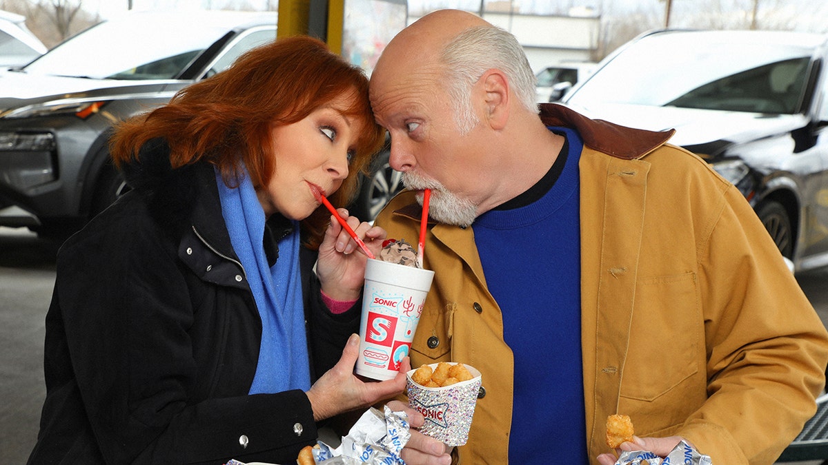 Reba McEntire in a black jacket and blue scarf and Rex Linn in a blue sweater and mustard jacket look into each other's eyes while drinking a Sonic shake