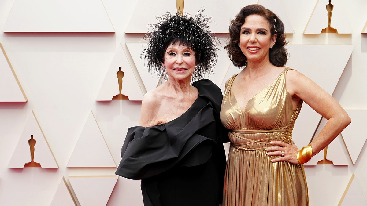 Rita Moreno in a black off the shoulder dress with a feathery hairpiece smiles on the carpet with daughter Fernanda in a gold gown at the Oscars