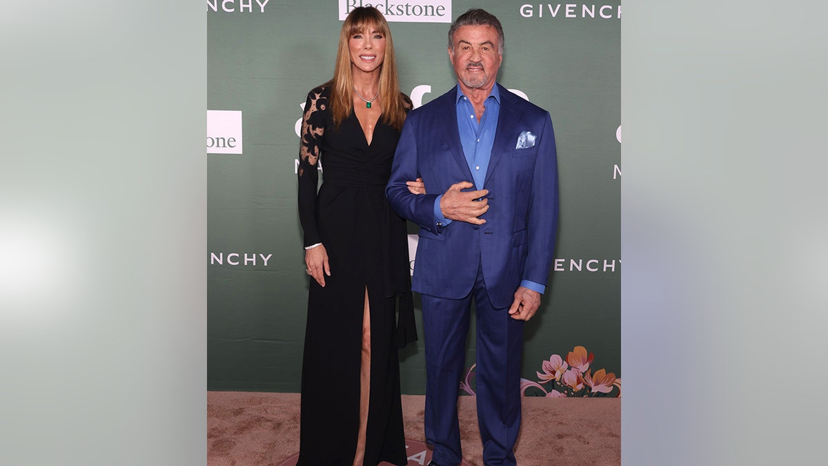 Jennifer Flavin in a black dress with a slit down the middle poses with husband Sylvester Stallone in a bright royal blue jacket