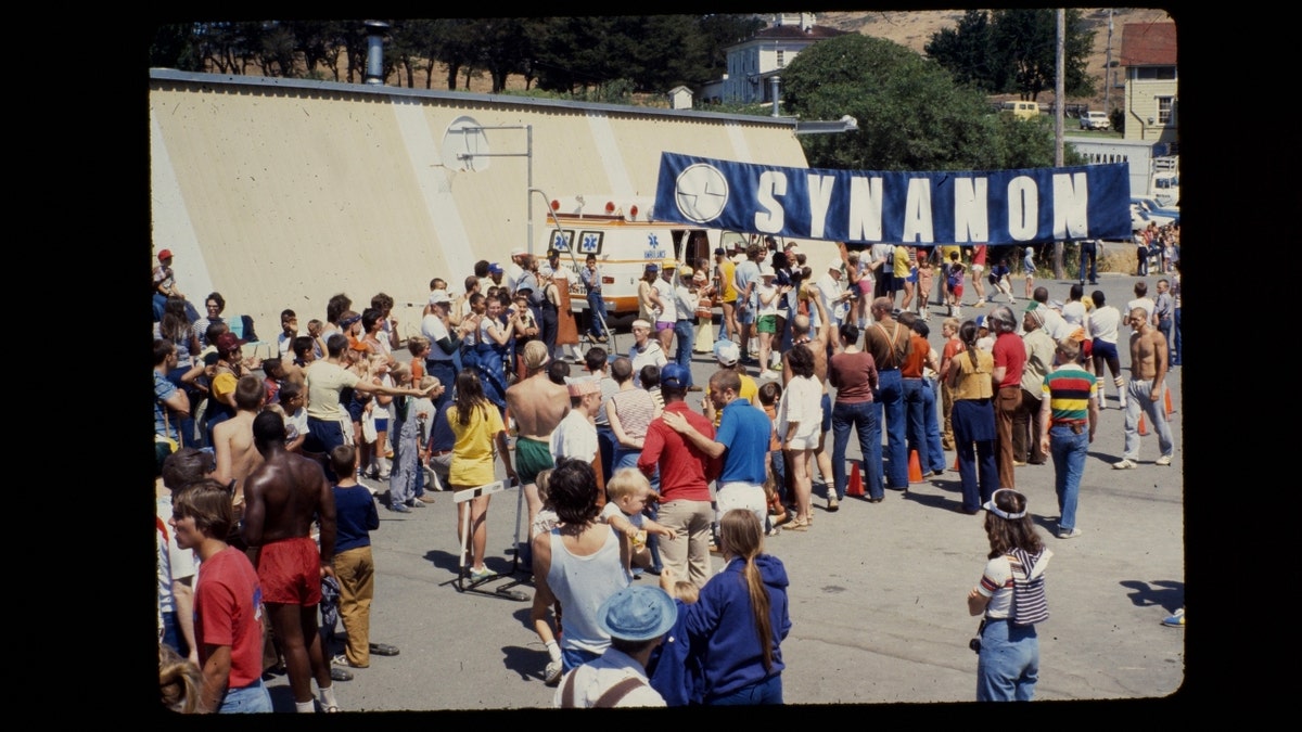 A crowd of Synanon members gather beneath a sign that says 'Synanon'