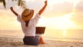 Work from anywhere. Rear view of young woman, female freelancer in straw hat working on laptop, keeping arms raised and cellebrating success while sitting on the tropical sandy beach at sunset