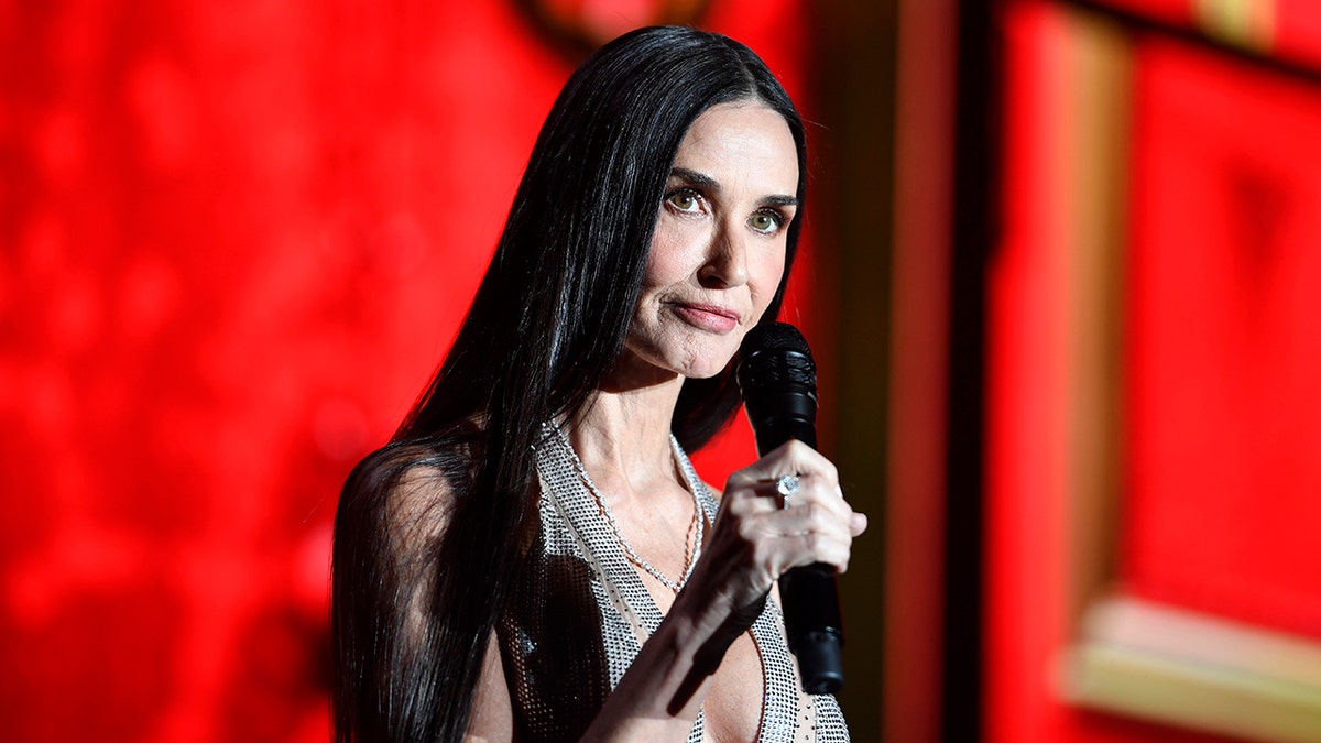 Demi Moore in a silver slinky dress holds a microphone and talks on stage