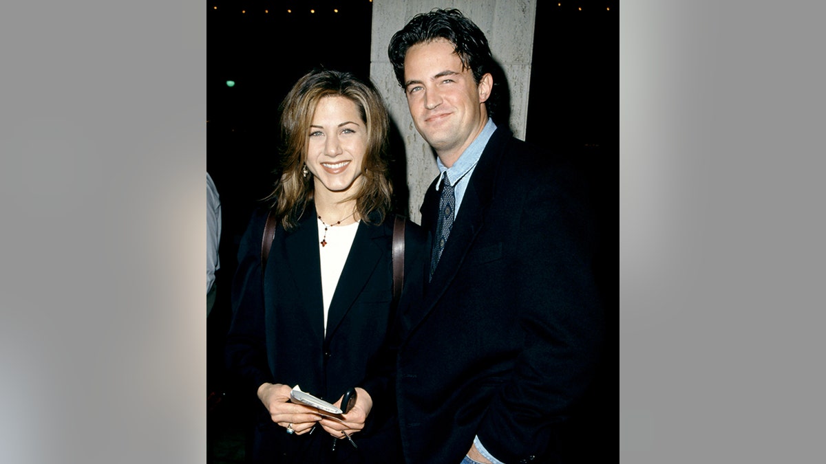 Jennifer Aniston in a black coat and white top smiles next to Matthew Perry