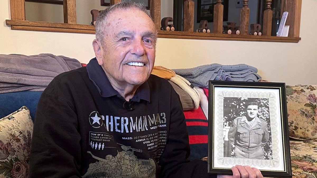 The 100-year-old Andy Negra Jr. smiles on a couch in his home as he holds up a black and white photograph of himself during his time with the Army's 128th Armored Field Artillery Battalion, 6th Armored Division.