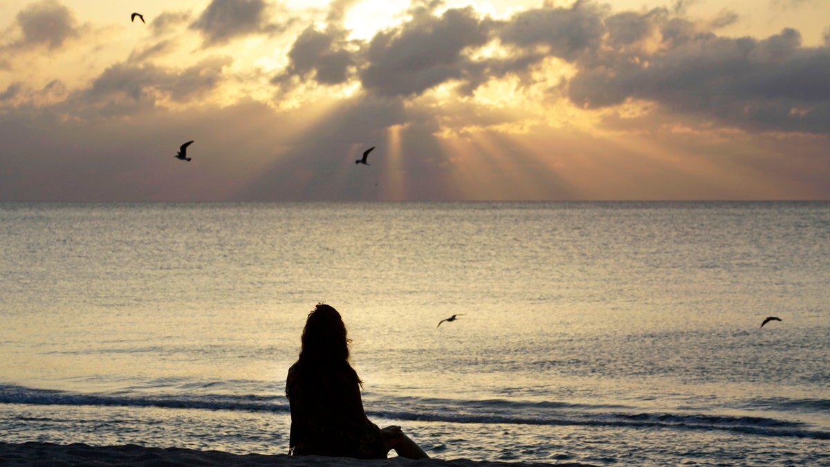 A woman meditates on the beach as the sun shines through the clouds