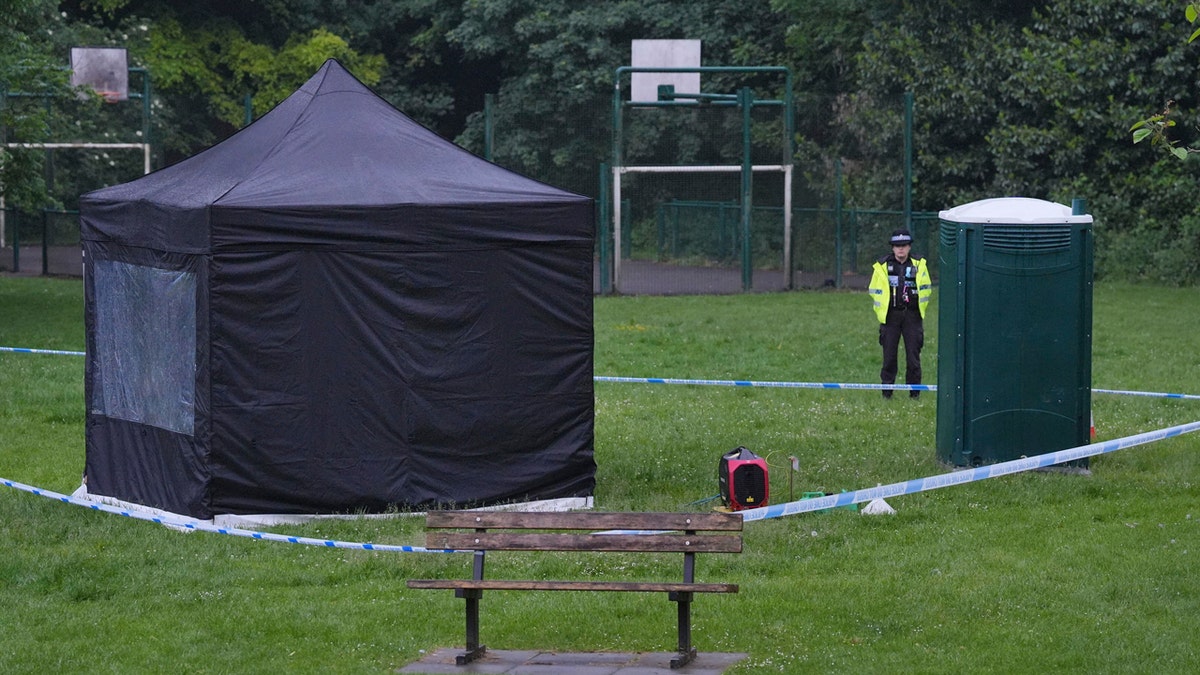 A police officer wearing a fluorescent yellow jacket stand next to a black forensic tent in Grenfell Park in Maidenhead, England, west of London, where Matthew Trickett was found dead.