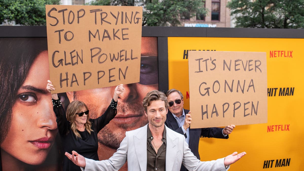 glen powell on red carpet lifting arms in front of parents holding signs