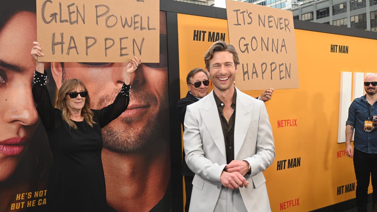 Glen Powell on the red carpet smiling in front of his parents with signs